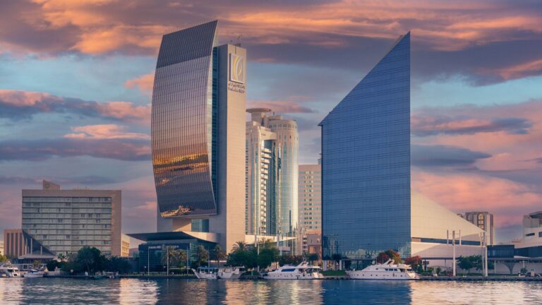 Glass office towers beside Dubai Creek catch the last of the sunset under a sky of pink and gold clouds, with small boats moored along the water.