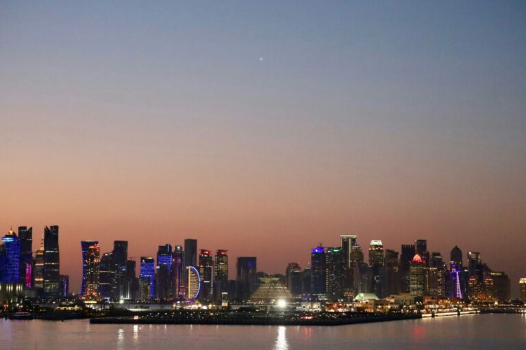 Doha's West Bay business district skyline at dusk, its office towers lit up under a pink and blue sky across calm water.